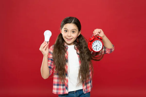 Czas świecenia. Pomysł i inspiracja. Inspirujące dziecko trzymać zegar czerwone tło. Inspiracja i burza mózgów. Energia elektryczna. Dziewczynka trzyma żarówkę. Słodkie dziecko szuka inspiracji — Zdjęcie stockowe