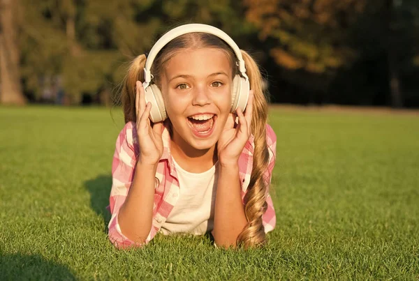 Menina pequena ouvir áudio livro. nova tecnologia para crianças. memórias felizes da infância. ouvir música. de volta à escola. O miúdo estuda no parque. relaxar na grama verde em fones de ouvido. viciado em aprendizagem. — Fotografia de Stock