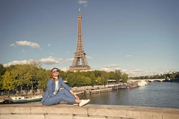 Uma mulher feliz senta-se no rio na Torre Eiffel em Paris, França. Mulher sorrir em óculos de sol no céu azul. Viajando em férias de verão. A sonhar com Paris. Menina com aparência de moda e beleza sensual — Fotografia de Stock