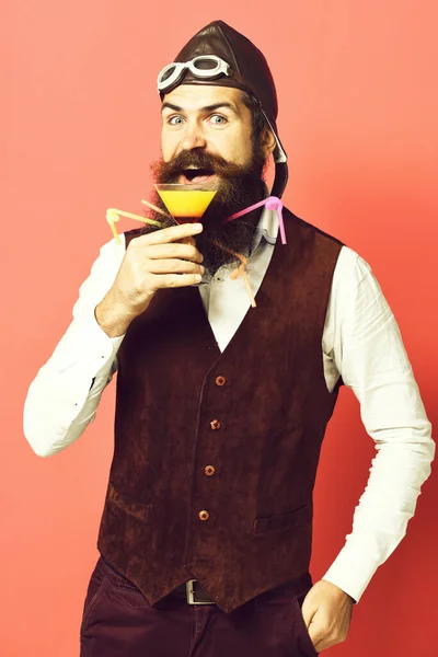 Bonito barbudo piloto ou aviador homem com longa barba e bigode no rosto feliz segurando vidro de bebida alcoólica em couro de camurça vintage colete com chapéu e óculos no fundo estúdio vermelho — Fotografia de Stock