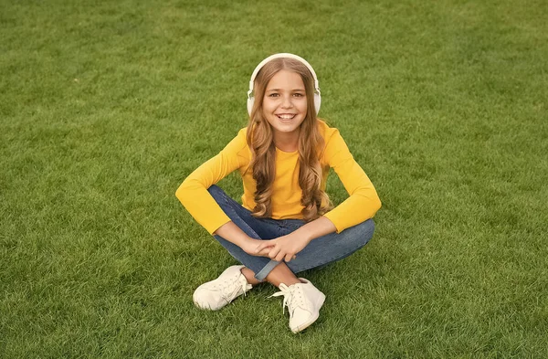 緑の芝生でリラックス。春のレジャータイム。幸せな子供時代。ヘッドセットの子供だ。幸せな女の子は好きな歌を聞く。子供の美しさとファッション。10代の女の子は屋外で音楽を聞く。自然界の音 — ストック写真
