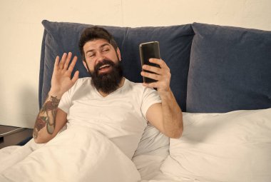 Herkes için çevrimiçi sohbet. Sakallı adam yatakta internette sohbet ediyor. Hippi el kameralı telefona el sallıyor. Çevrimiçi toplantı. Mobil cihazda video konferansı. Çevrimiçi video görüşmesi. Akıllı telefon iletişimi