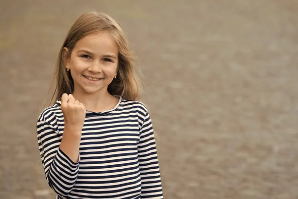 Poder femenino. Chica feliz mostrar puño al aire libre. Belleza mirada de niña. Un niño pequeño sonríe con un top a rayas. Estilo casual. Moda de verano. Armario de moda. Peluquería. Impresionante y magnífico, espacio de copia — Foto de Stock