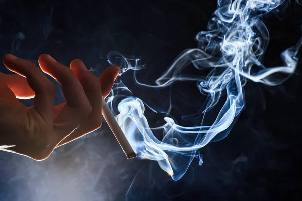 男性手握燃烧的香烟，卷起烟幕，黑黑的背景，吸烟的习惯 — 图库照片
