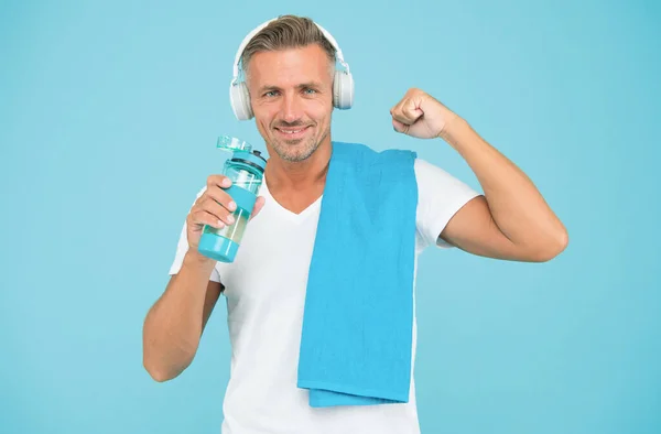 スポーツマンは健康的なライフスタイルを促進する。青い背景に幸せなスポーツマン。スポーツマンにタオルを合わせなさい。ハンサムなスポーツマンは腕の筋肉を曲げます。訓練中に水を飲む。音楽を聴くこととのワークアウト — ストック写真