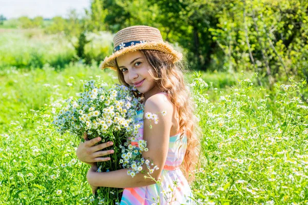 Happy little kid wear sun hat on long wavy hair holding beautiful romomile flowers spa and beauty care on sunny summer landscape, look — стоковое фото