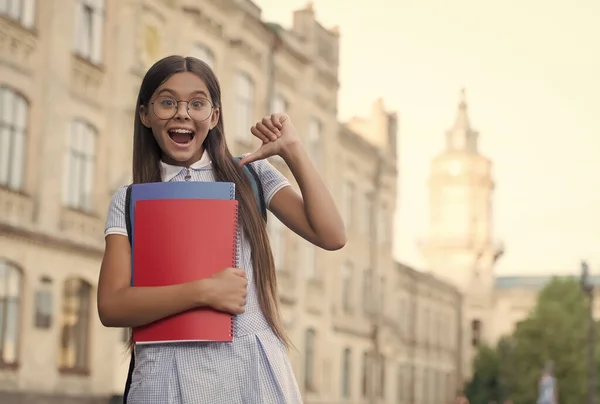 Szczęśliwe małe dziecko okulary girlin wskazując palcem kciuka w książkach szkolnych na zewnątrz, informacje, przestrzeń kopiowania — Zdjęcie stockowe