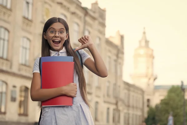 Gafas de niña pequeña y feliz apuntando con el dedo pulgar a los libros escolares al aire libre, información, espacio para copiar — Foto de Stock