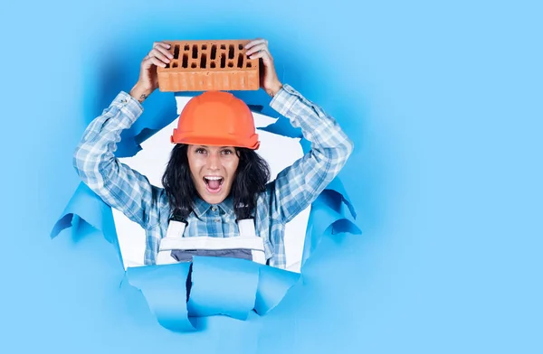 Machinist houdt Brick vast. Jonge werkneemster op blauwe achtergrond. reparatie en renovatie. vrouw draagt een beschermende helm. Meisjesbouwer met harde hoed op de bouwplaats. plaats voor kopieerruimte. — Stockfoto