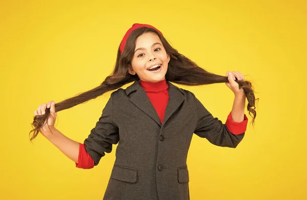 Gelukkig klein kind in schooluniform houden lang brunette haarlokken gele achtergrond, salon — Stockfoto
