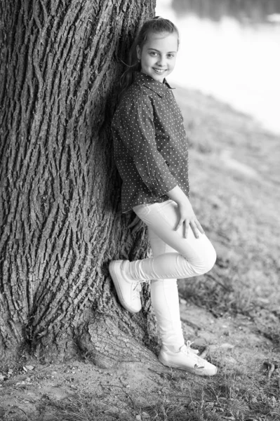 Desfrutando de férias de verão. Menina pequena relaxando na árvore no dia de verão. Criança adorável com longos cabelos loiros em estilo casual na paisagem de verão. Criança vestindo roupas de verão de moda — Fotografia de Stock