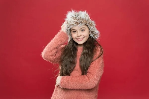 Jag känner mig så mysig. Barn lång hår mjuk hatt. Vintermode koncept. Varm mössa för kallt vinterväder. Ung flicka leende röd bakgrund. Mjukt lurvigt tillbehör. Vintersäsong. Liten fashionista koncept — Stockfoto