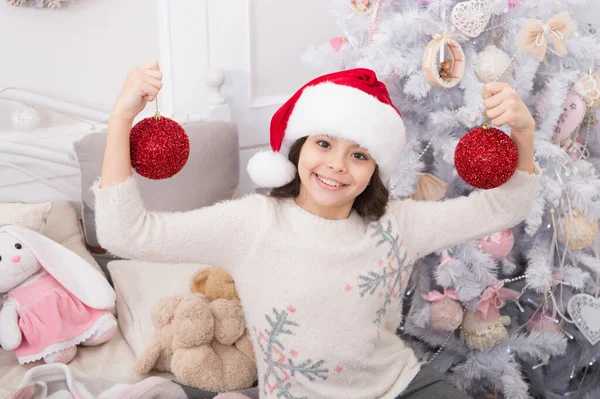 Playful mood. Cheerful kid. Creating festive atmosphere. Child decorating christmas tree with red ball. Girl kid decorating christmas tree. Kid in santa hat decorating christmas tree. Good vibes — Stock Photo, Image