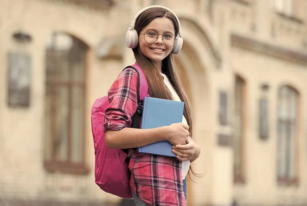 Glückliches Kind hält Schulbuch in der Hand und hört Audio-Podcast in modernen Kopfhörern im Freien, Musikbibliothek — Stockfoto