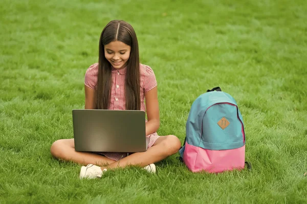 Bambino felice utilizzando il computer portatile per l'educazione scolastica online seduto nel parco su erba verde con zaino, infanzia — Foto Stock