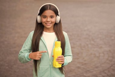 Yüzünde kulaklıkla gülümseyen liseli kız plastik şişeyi işaret ediyor açık havada su içiyor, fotokopi çekiyor, susamış.