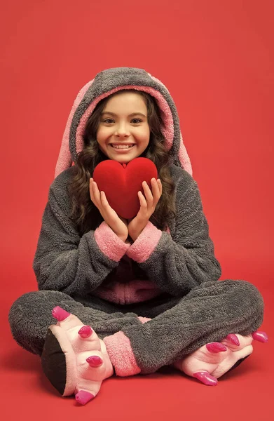 Happy kid hold big red valentines heart sitting in cosy pajamas, cardiovascular