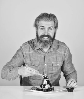 Doğu kültürü. Sakallı hippi rulo yiyor. Japon yemek teslimatı. Suşi ruloları. Asya yemeği. Suşi siparişi. Adam evde suşi yiyor. Çubuklu suşi ve zencefil. Japon mutfağı.