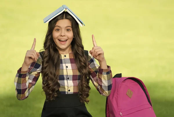 Happy small kid in school uniform hold book on head pointing index fingers up sunny outdoors, idea