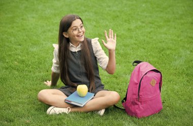 Okul üniformalı mutlu çocuk jest yapıyor. Yeşil çimlerde kitap ve elmayla rahatlıyor. Eğitim.