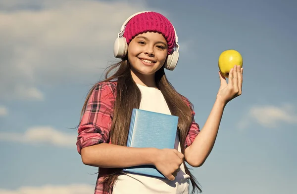 Criança feliz em estilo casual segurar maçã e livro escolar ouvir áudio em fones de ouvido, conhecimento — Fotografia de Stock