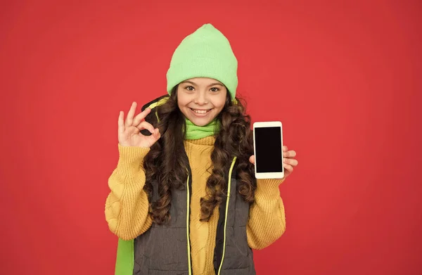 Стильная девочка в повседневной теплой одежде показать хорошо жест для хорошего качества смартфона, совершенство — стоковое фото