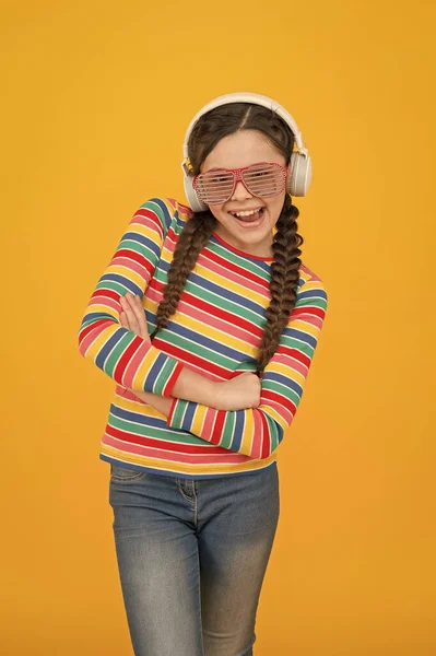Glücklich Teenager-Mädchen mit geflochtenen Haaren tragen Regenbogenfarbe und Glamour-Party-Brille hören Musik in Kopfhörer auf gelbem Hintergrund, Technologie — Stockfoto