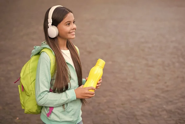 Hidratação de saúde para criança com garrafa de água ouvindo música em fones de ouvido e levar mochila escolar ao ar livre, espaço de cópia, infância — Fotografia de Stock