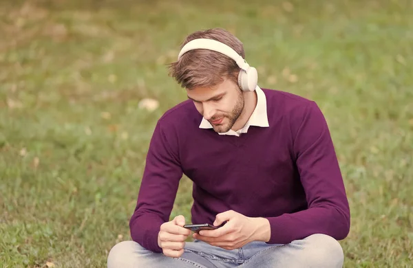 녹색 잔디에 있는 잘생긴 남자 휴대 전화 mp3 플레이어에서 노래를 바꾸고 디지탈 시대의 이어폰으로 음악을 듣습니다. — 스톡 사진