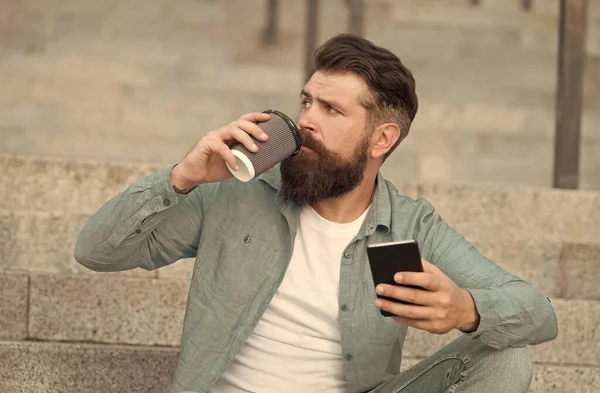 Hombre barbudo guapo relajante teléfono móvil y taza de café. Vivir la vida en línea. Redes sociales. Comunicación en línea de la vida moderna. Tipo con fondo de espacio urbano smartphone. Tecnología moderna — Foto de Stock