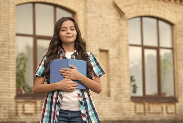 Симпатичный мечтательный ребенок с длинными волосами в непринужденном стиле держит школьную книгу закрытыми на школьном дворе, мечтая — стоковое фото