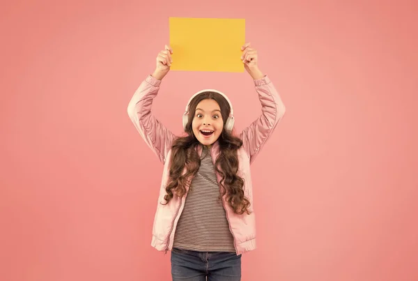 Fröhliches Teenie-Mädchen hört Musik mit Kopfhörern und präsentiert leeres Papier, zurück in die Schule — Stockfoto