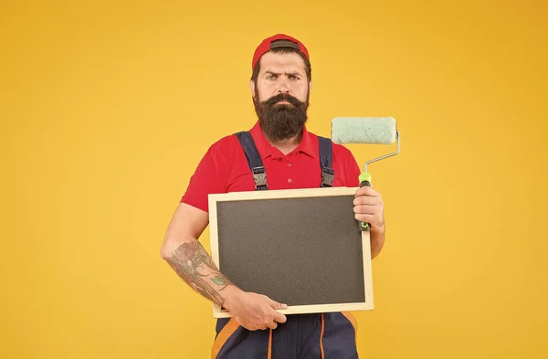 Ältere bärtige Kerl Maler Arbeiter in Kesselanzug Uniform mit Farbroller Werkzeug zum Streichen der Wand und halten Tafel für Kopierraum Werbung, Werbung — Stockfoto