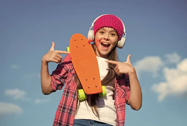 信頼できるスケートボードだ。幸せな女の子はペニーボードを指している。青い空にジェスチャーを指している小さなスケーター。広告のためのポイント。マーケティングとプロモーション。輸送、コピースペースに適しています — ストック写真