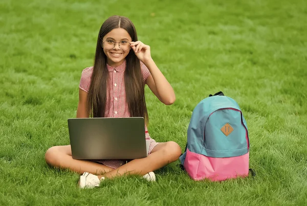 Teen girl utilizzare il computer su erba verde nel parco. bambino con zaino e taccuino. nuove tecnologie nella vita moderna. studio. bambino felice in occhiali che lavora su computer portatile. educazione online. torna a scuola — Foto Stock