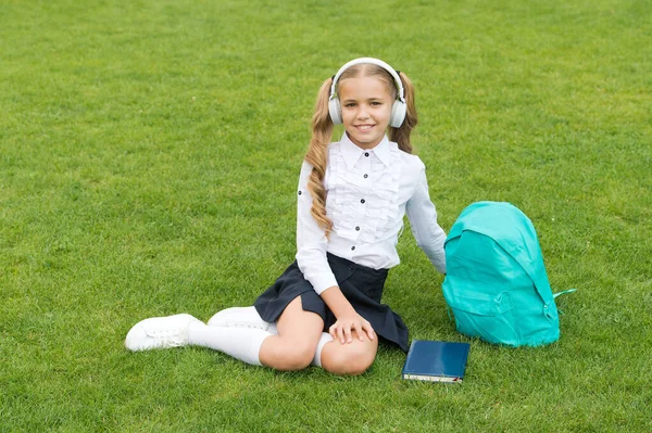 Happy schoolchild v uniformě poslouchat hudbu ve sluchátkách sedí na zelené trávě, školní prázdniny — Stock fotografie