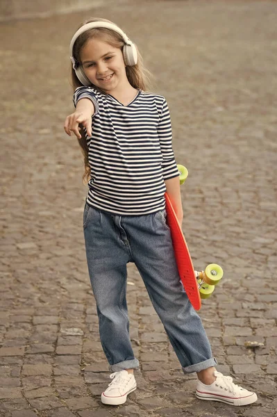 Criança feliz apontar dedo segurando penny skate e ouvir música em fones de ouvido modernos na rua de verão, ao ar livre — Fotografia de Stock