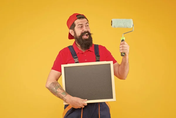 Mutlu sakallı, kazan dairesinde çalışan bir boyacı duvarları boyamak için boya silindiri kullanıyor ve reklam için karatahta tutuyor. — Stok fotoğraf