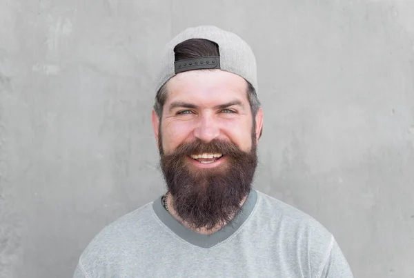 Garder les cheveux en bonne santé. Joyeux hipster avec barbe en forme et coiffure coiffée portant une casquette de baseball. Homme barbu souriant avec des poils de visage non rasés. Poilu avec barbe élégante et les cheveux de moustache sur le mur gris — Photo