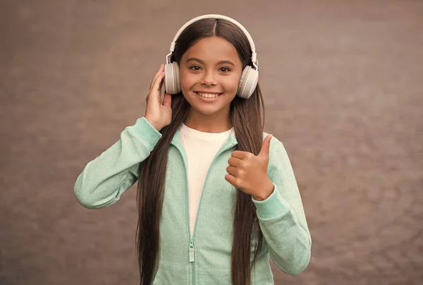 Happy smiling child enjoys listens to music in headphones. kid listen song outdoor. walking with favorite tune. education online concept. childhood happiness. elearning of student girl. thumb up — Stock Photo, Image