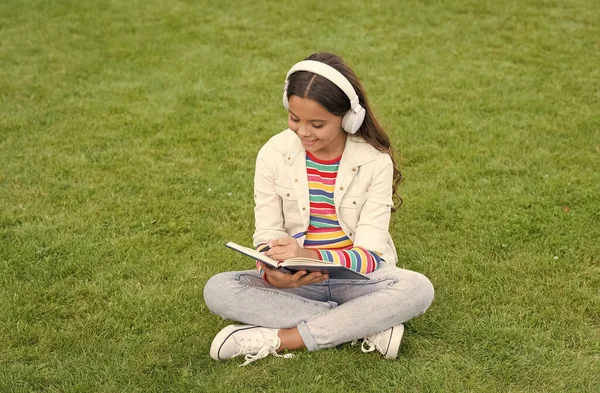 写歌歌词。快乐童年的概念。孩子把记忆写在日记里。女学生拿着笔记本放松。快乐的孩子在耳机里听音乐。在绿草上的小女孩。孩子们喜欢公园自然的歌声 — 图库照片