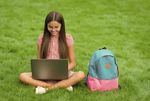 Intelligente bambino felice indossare gli occhiali utilizzando il computer portatile per fare i compiti scolastici online seduto nel parco su erba verde con zaino, educazione — Foto Stock