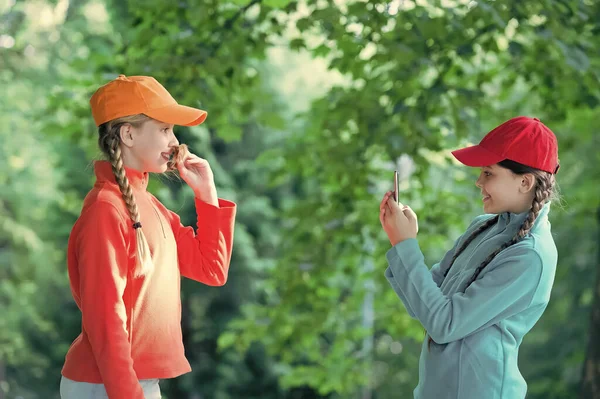 Zorgeloze kinderen poseren buiten voor foto. Positieve modellen. Casual hipsters outfit. portret van gelukkige zussen met smartphone. Veel plezier, kleine kinderen. glimlachende hippe meisjes in sportkleding. Hulpzaam instrument — Stockfoto