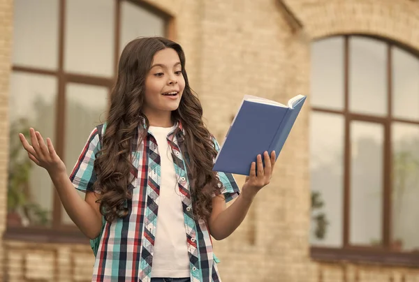Счастливый ребенок с длинными волнистыми волосами в непринужденном стиле читал библиотечную книгу, готовясь к школьному уроку на открытом воздухе, литературу — стоковое фото