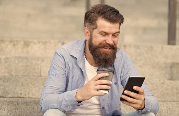Comunicación en línea de la vida moderna. Tipo con fondo de espacio urbano smartphone. Hombre barbudo guapo relajante teléfono móvil y taza de café. Tecnología moderna. Vivir la vida en línea. Redes sociales — Foto de Stock