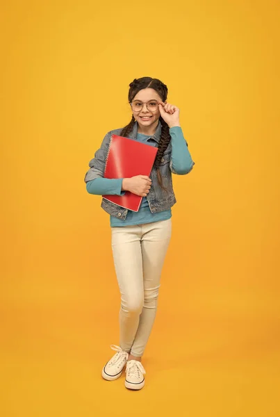 Glimlachend kind meisje met stijlvol gevlochten haar dragen bril houden werkboek op gele achtergrond, terug naar school — Stockfoto