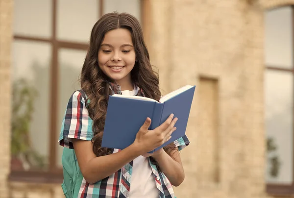 Glückliches Kind mit langen welligen Haaren in lässigem Stil lesen Schulbuch auf dem Schulhof, lesen — Stockfoto
