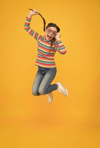 Niña saltando feliz con el pelo trenzado usar el color del arco iris y gafas de fiesta glamour sobre fondo amarillo, extático — Foto de Stock