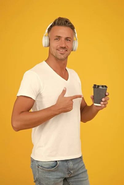 Homem feliz ouvir música em fones de ouvido modernos apontando dedo em takeaway café copo fundo amarelo, ordem — Fotografia de Stock
