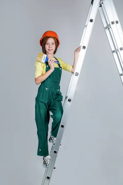 Teen girl laborer in protective helmet and uniform on ladder with painting brush, painter — Stock Photo, Image